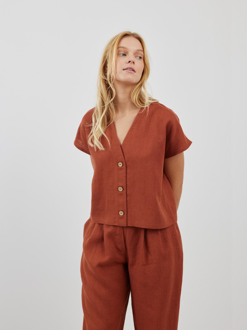 May include: A rust-colored linen top with a v-neck and button closure. The top has short sleeves and is paired with matching rust-colored linen pants.