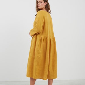 May include: A long, mustard yellow linen dress with a gathered skirt and long sleeves. The dress is loose-fitting and has a simple, casual style.