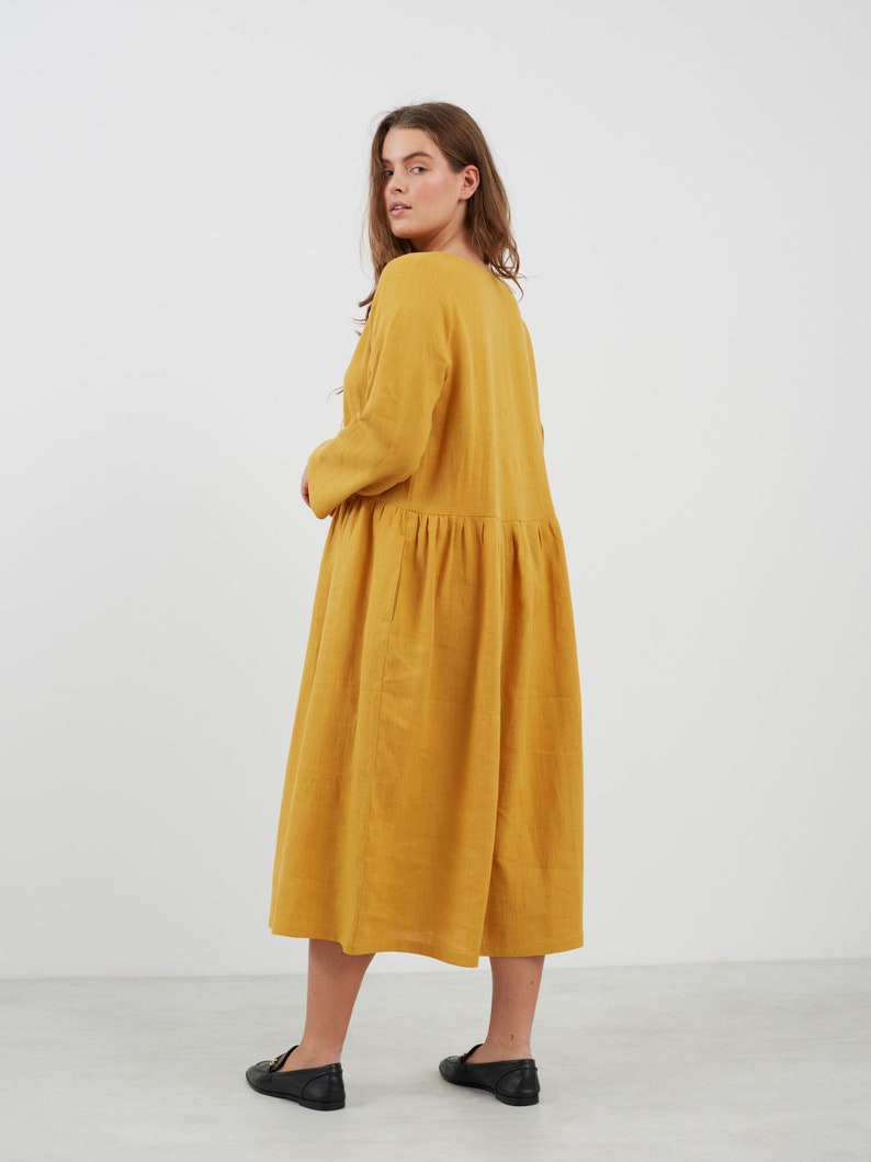 May include: A long, mustard yellow linen dress with a gathered skirt and long sleeves. The dress is loose-fitting and has a simple, casual style.