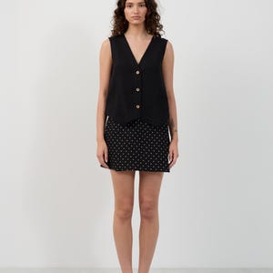 May include: A woman wearing a black sleeveless top with wooden buttons and a black skirt with white polka dots. She is also wearing black loafers with gold accents. The outfit is simple and stylish.
