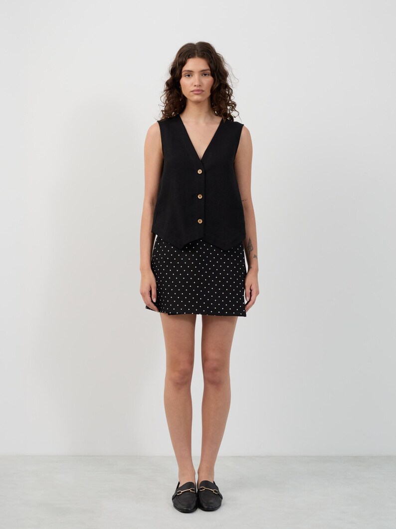 May include: A woman wearing a black sleeveless top with wooden buttons and a black skirt with white polka dots. She is also wearing black loafers with gold accents. The outfit is simple and stylish.