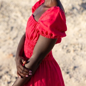 May include: A woman wearing a red, sleeveless dress with a square neckline and puffed sleeves. The dress has a gathered waist and a flowing skirt.