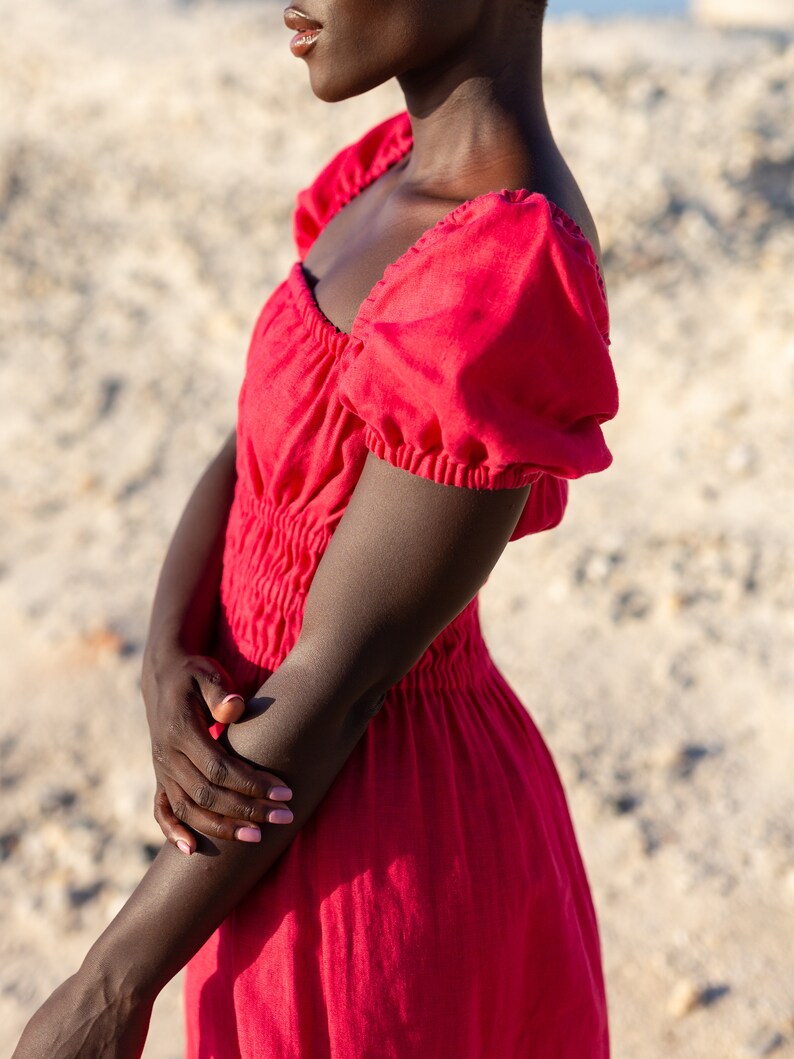 May include: A woman wearing a red, sleeveless dress with a square neckline and puffed sleeves. The dress has a gathered waist and a flowing skirt.