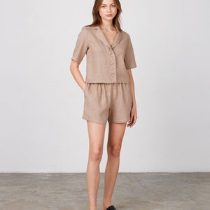 May include: A woman is wearing a light brown linen short-sleeved shirt and matching shorts. She is standing on a concrete floor and wearing black slip-on shoes.