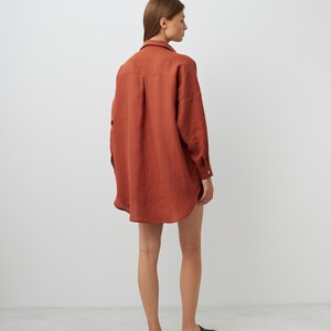 May include: A rust-colored linen shirt with long sleeves and a relaxed fit. The shirt is worn by a person and is shown from the back.