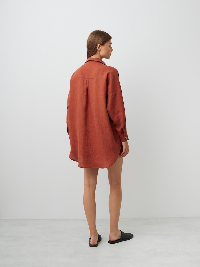 May include: A rust-colored linen shirt with long sleeves and a relaxed fit. The shirt is worn by a person and is shown from the back.