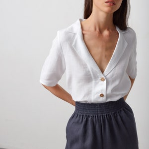 May include: A white linen shirt with a collar and a button-up front. The shirt has short sleeves and a relaxed fit. The model is wearing the shirt with dark gray pants.