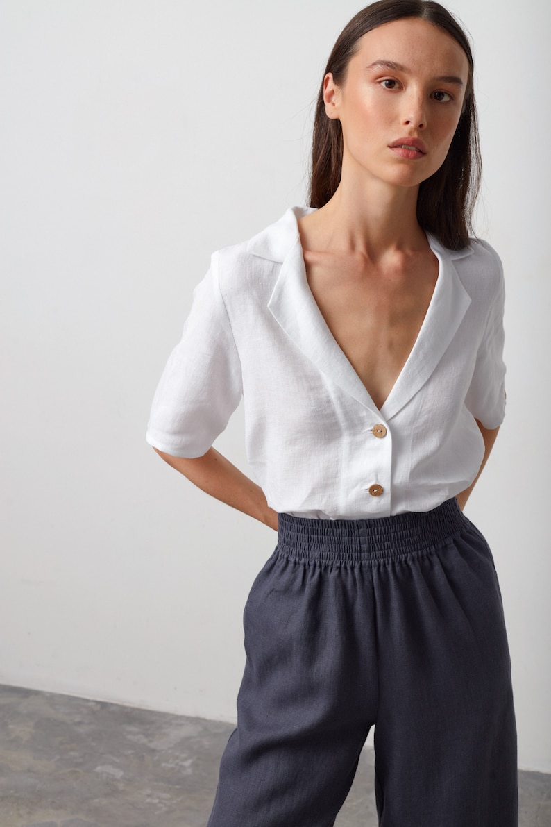 May include: A white linen shirt with a collar and a button-up front. The shirt has short sleeves and a relaxed fit. The model is wearing the shirt with dark gray pants.