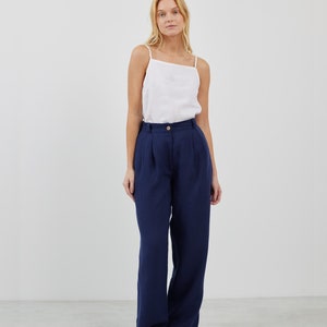 May include: A woman is wearing a white tank top and dark blue wide-leg pants. She is standing on a light gray floor.