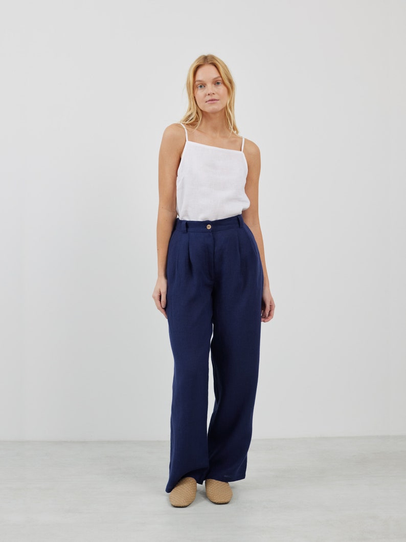 May include: A woman is wearing a white tank top and dark blue wide-leg pants. She is standing on a light gray floor.