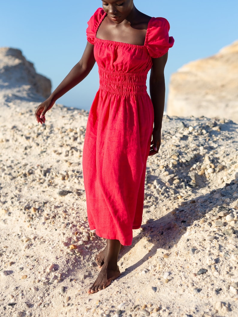 May include: A woman wearing a bright red, linen dress with puffed sleeves and a fitted bodice. The dress has a square neckline and a gathered waist. The woman is walking on a sandy beach.