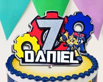May include: A yellow and blue Transformers themed birthday cake with a silver number 7 and the name "DANIEL" on top. The cake is decorated with blue frosting and yellow sprinkles.