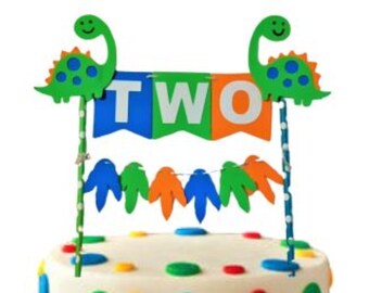 May include: A white cake decorated with colorful polka dots and topped with a dinosaur cake topper. The topper features two green dinosaurs holding a banner that says "TWO".