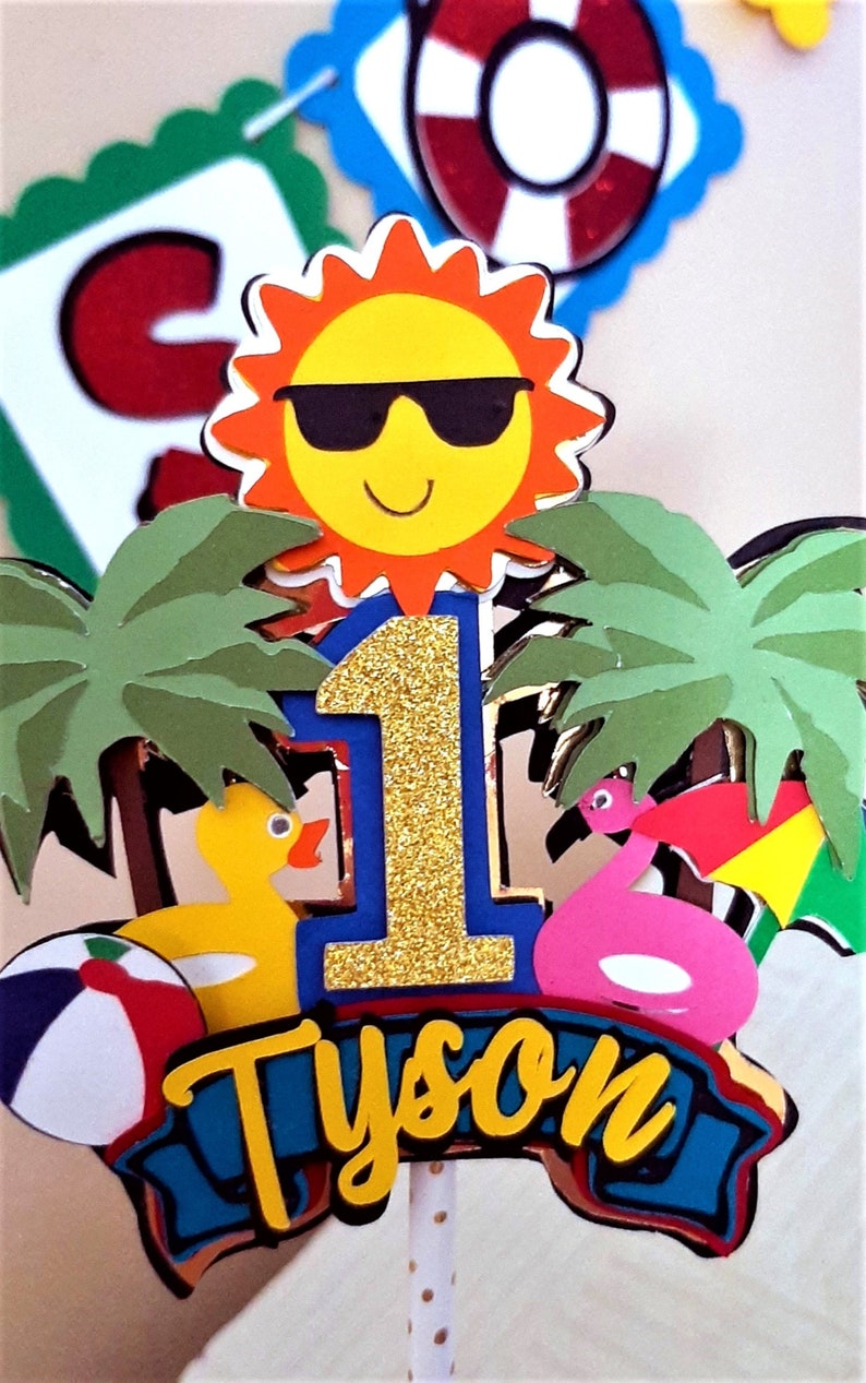 May include: A tropical-themed cake topper with a yellow sun wearing sunglasses, palm trees, a blue number one with gold glitter, a yellow rubber duck, a pink flamingo, a beach ball, and a blue banner with the name "Tyson" in yellow letters.