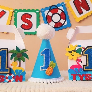 May include: Two blue and white favor boxes with a sun, palm tree, flamingo, and rubber ducky design. The boxes say "Tyson" and have the number "1" on them. A blue party hat with a white pom-pom on top has the number "1" and a pineapple on it.