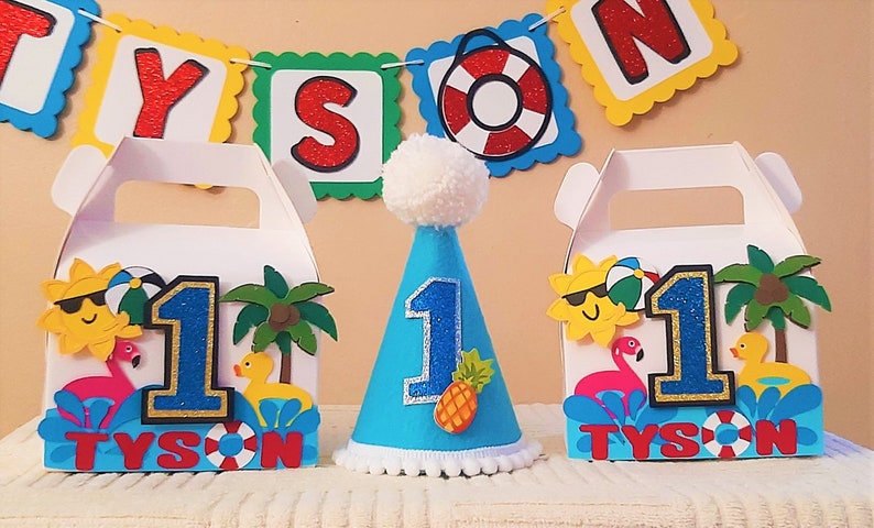 May include: Two blue and white favor boxes with a sun, palm tree, flamingo, and rubber ducky design. The boxes say "Tyson" and have the number "1" on them. A blue party hat with a white pom-pom on top has the number "1" and a pineapple on it.