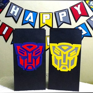May include: Two black paper bags with Transformers logos. The bag on the left has a red and blue Autobot logo. The bag on the right has a yellow and gray Decepticon logo. The bags are decorated with a Happy Birthday banner.