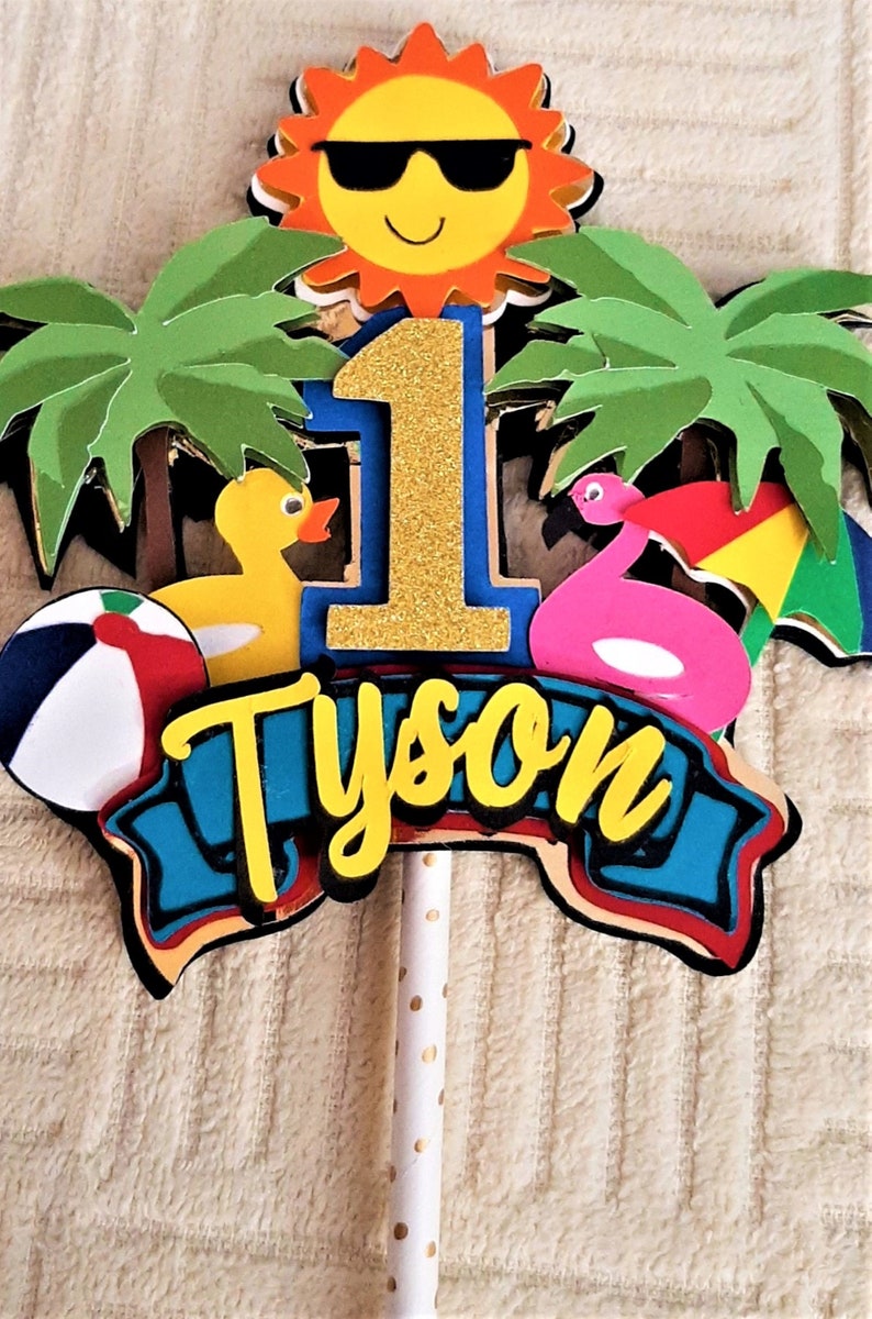 May include: A cake topper featuring a yellow sun wearing sunglasses, palm trees, a pink flamingo float, a yellow rubber duck, a beach ball, a blue banner with the name "Tyson" in yellow letters, and a gold glitter number "1".
