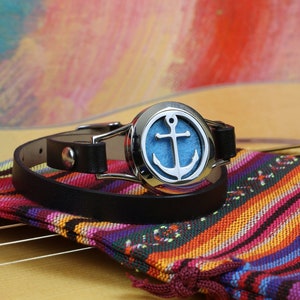 May include: A black leather bracelet with a silver diffuser charm featuring a blue anchor. The bracelet is on a colorful striped fabric pouch.