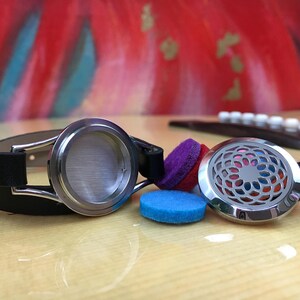 May include: A black leather bracelet with a silver essential oil diffuser charm. The charm has a decorative design and is shown with three colourful felt pads.