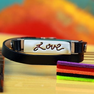 May include: A black leather bracelet with a silver rectangular charm that says "Love" in cursive. The bracelet is lying on a wooden surface with a stack of colorful felt pads.