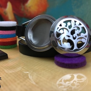 Pode incluir: Um bracelete difusor de metal prateado com um design de árvore. O bracelete está aberto e a almofada difusora é visível. A almofada difusora é roxa.