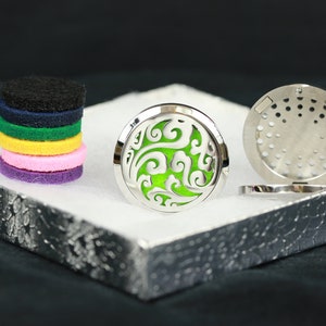 Könnte beinhalten: Ein silberner Diffusor-Anhänger für ätherische Öle mit einem grünen floralen Design. Der Anhänger ist geöffnet und die Rückseite ist sichtbar. Ein Stapel bunter Filzpads ist ebenfalls zu sehen.