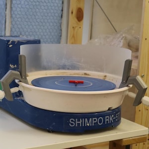 May include: A blue and white pottery wheel with a clear plastic splash guard. The wheel is labeled "SHIMPO RK-5T".