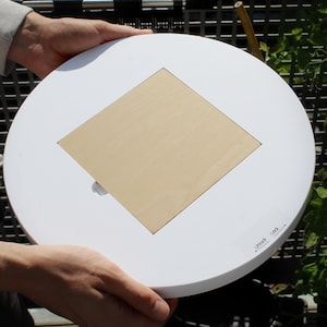 May include: A white, round object with a square, light-colored wooden insert in the center. The object has the words "Unlock" and "Lock" printed on the surface. The object is being held by a person.