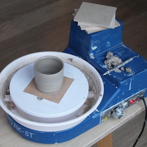 May include: A blue pottery wheel with a white ceramic plate and a clay pot on top. The wheel has a control panel with a red light and a power cord. The text "ORK-5T" is visible on the wheel.