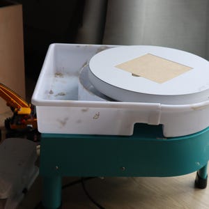 May include: A white pottery wheel with a teal base and a white platform. The platform has a small wooden square in the center.