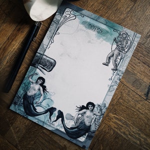 May include: A white and brown nautilus shell sits on a wooden surface next to a blue and white notepad with a mermaid and a diver illustration. The notepad has the word "notes" written on it.