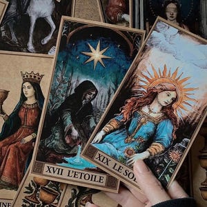 Op de afbeelding: Een verzameling tarotkaarten met vintage illustraties. De kaarten tonen afbeeldingen van figuren, sterren en hemellichamen. De kaarten zijn in verschillende kleuren, waaronder blauw, goud en rood. De kaarten worden door een persoon vastgehouden.