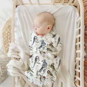 Peut inclure: Un bébé endormi enveloppé dans une couverture blanche ornée d'un motif d'oiseaux colorés et de feuillage vert. Les oiseaux sont dans des tons de bleu, vert et marron. Le bébé est dans un berceau en bois avec un drap blanc.