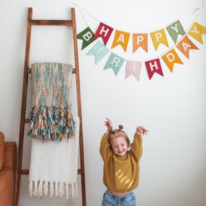 May include: A young child wearing a yellow sweater and blue jeans stands in front of a white wall with a colorful "Happy Birthday" banner hanging above.