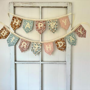 May include: A two-tiered fabric bunting banner featuring triangular pennants in various floral patterns of blue, pink, and brown. Each pennant displays a cream-colored letter, collectively spelling out the phrase HAPPY BIRTHDAY in white.