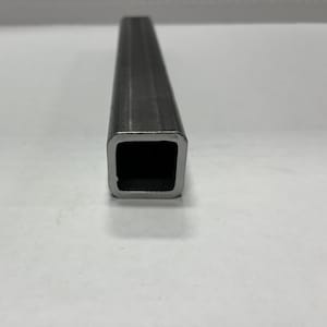 May include: A close-up view of a square metal tube. The tube is made of a dark gray metal and has a hollow, square opening. The edges of the tube are slightly rounded. The background is a plain white surface.