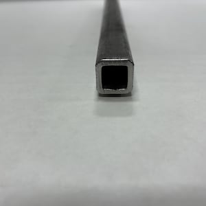 May include: Close-up of a square metal tube with a hollow interior. The tube is a dull silver color and appears to be made of steel or a similar metal. The end of the tube is in sharp focus, showing the square opening.