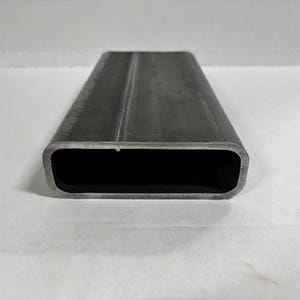 May include: A rectangular metal tube with rounded edges. The tube is made of a dark gray metal and has a seam running down the center. The interior of the tube is black. The tube is set against a white background.