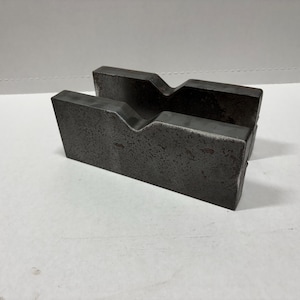 May include: Two dark gray metal blocks with a V-shaped groove cut into the top. The blocks are rectangular and have a rough, textured surface. The objects are likely used for industrial or mechanical purposes.