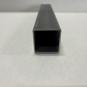May include: A close-up of a rectangular, hollow metal tube. The tube is a dark gray color and has a square opening. The metal appears to be unfinished or raw. The tube is lying on a white surface.