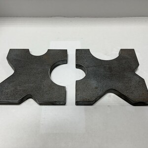 May include: Two metal pieces with a unique shape, each with a cut-out circle in the center. The pieces are gray and have a rough texture.