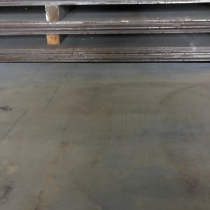 May include: A close-up of a large, flat, gray metal sheet. The sheet has a rough, textured surface with visible scratches and marks. The sheet is lying on a flat surface.
