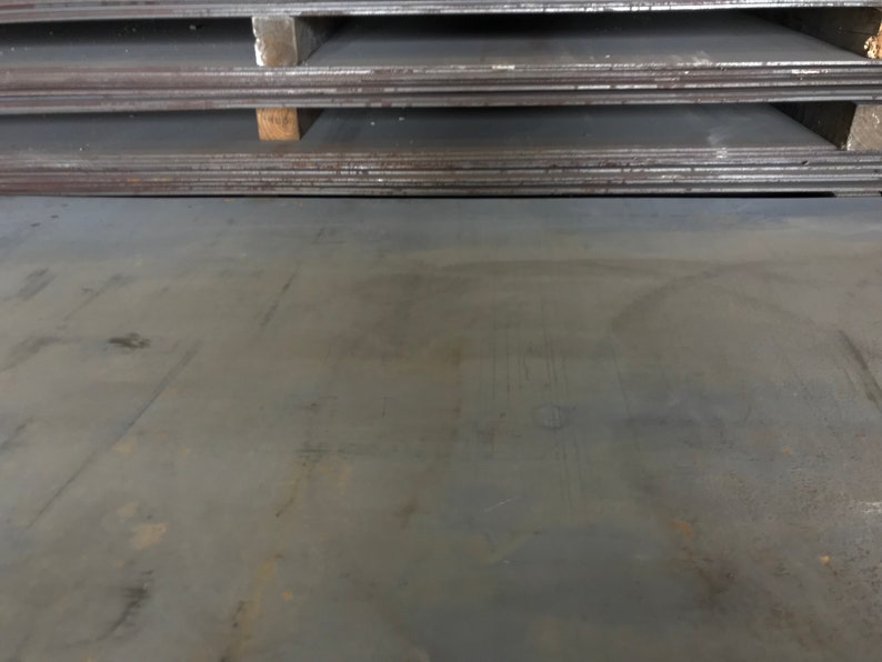 May include: A close-up of a large, flat, gray metal sheet. The sheet has a rough, textured surface with visible scratches and marks. The sheet is lying on a flat surface.