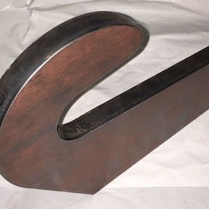 May include: A large, abstract metal sculpture with a curved, letter-like shape. The sculpture is made of thick, dark brown metal with a contrasting dark edge. The overall design is modern and industrial.