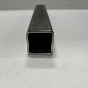 May include: A close-up view of a rectangular, hollow metal tube. The tube is a dark gray color and has a square opening. The edges of the tube are slightly rounded. The background is a plain white surface.