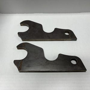 May include: Two metal brackets with a rounded hole and a smaller hole. The brackets are made of raw metal and have a rough, unfinished look.