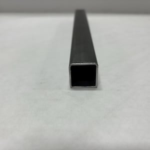 May include: A close-up shot of a dark gray, square metal tube. The tube has a hollow, black interior and a matte finish. The metal appears to be unfinished or raw. The tube is positioned vertically.