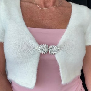 Könnte beinhalten: Weiße, flauschige, verkürzte Strickjacke mit einem Verschluss aus Kristallen.