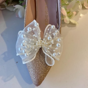 May include: A pointed-toe shoe with a gold glitter finish, featuring a large, sheer white organza bow attached to the front. The bow is embellished with numerous small, round white faux pearls of varying sizes scattered across the fabric.